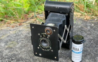 Titelbild zum Beitrag. Neben der Vest Pocket Autographic Kamera steht eine Filmdose mit einem 127er Rollfilm