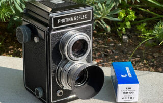 Die Photina Reflex III von Photavit steht auf einer Mauer. Daneben liegt ein Rollfilm Kentmere 100