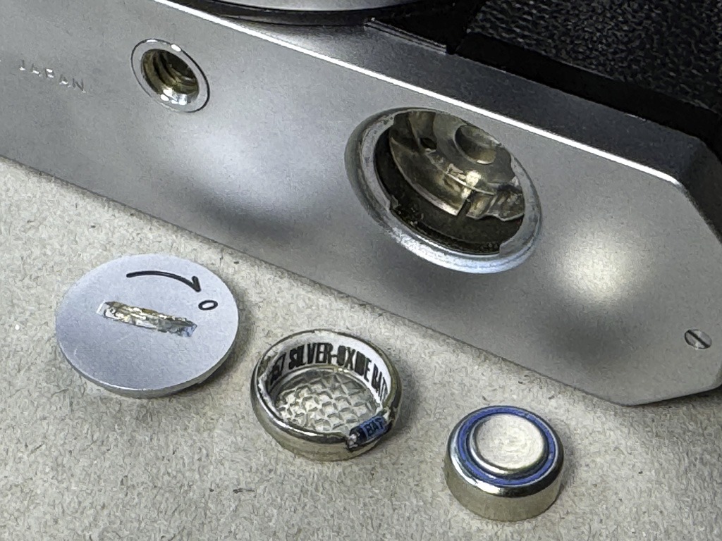 Das geöffnete Batteriefach der Nikkormat FT. Davor liegen eine SR44-Batterie, der benötigte Adapter und der Deckel des Batteriefachs.